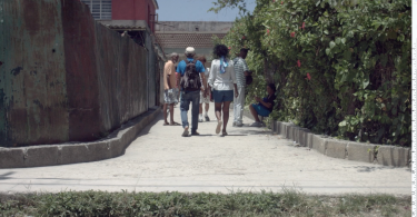 Zaima y Tomás caminan juntos por un pasillo estrecho en su barrio, rodeados de otras personas, vegetación y estructuras deterioradas.